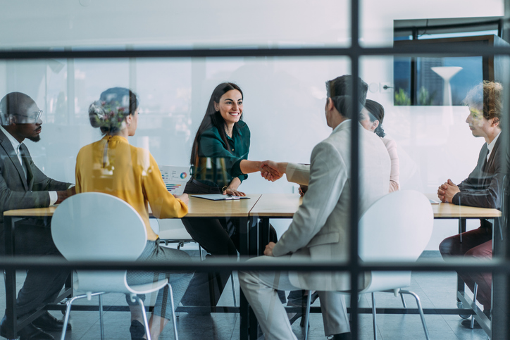 Business people from mixed ethic backgrounds, meeting and shaking hands around board room table.
